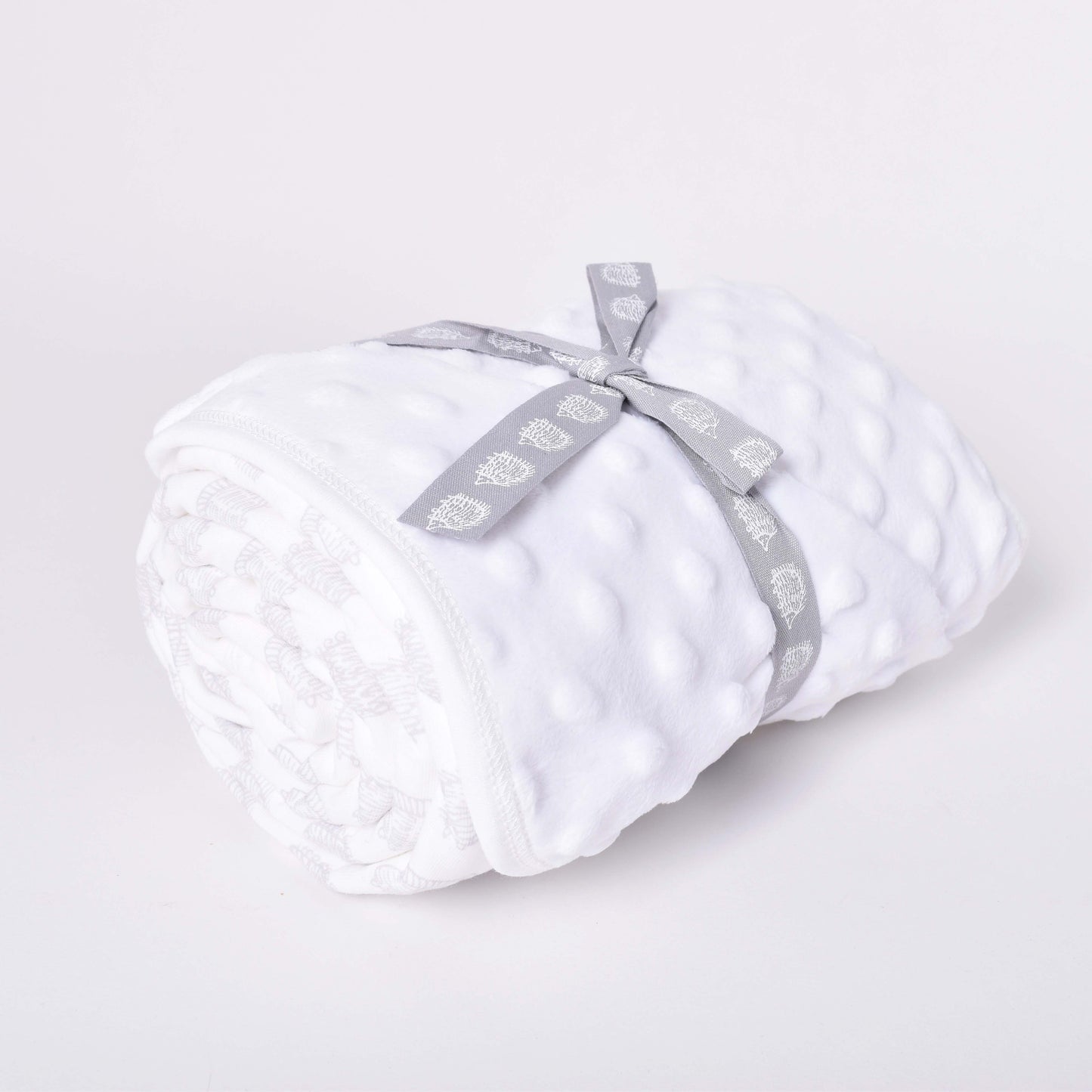 Vit babyfilt med motiv av igelkottar i mönsterdesign av Lisa Larson, hoprullad med grå rosett