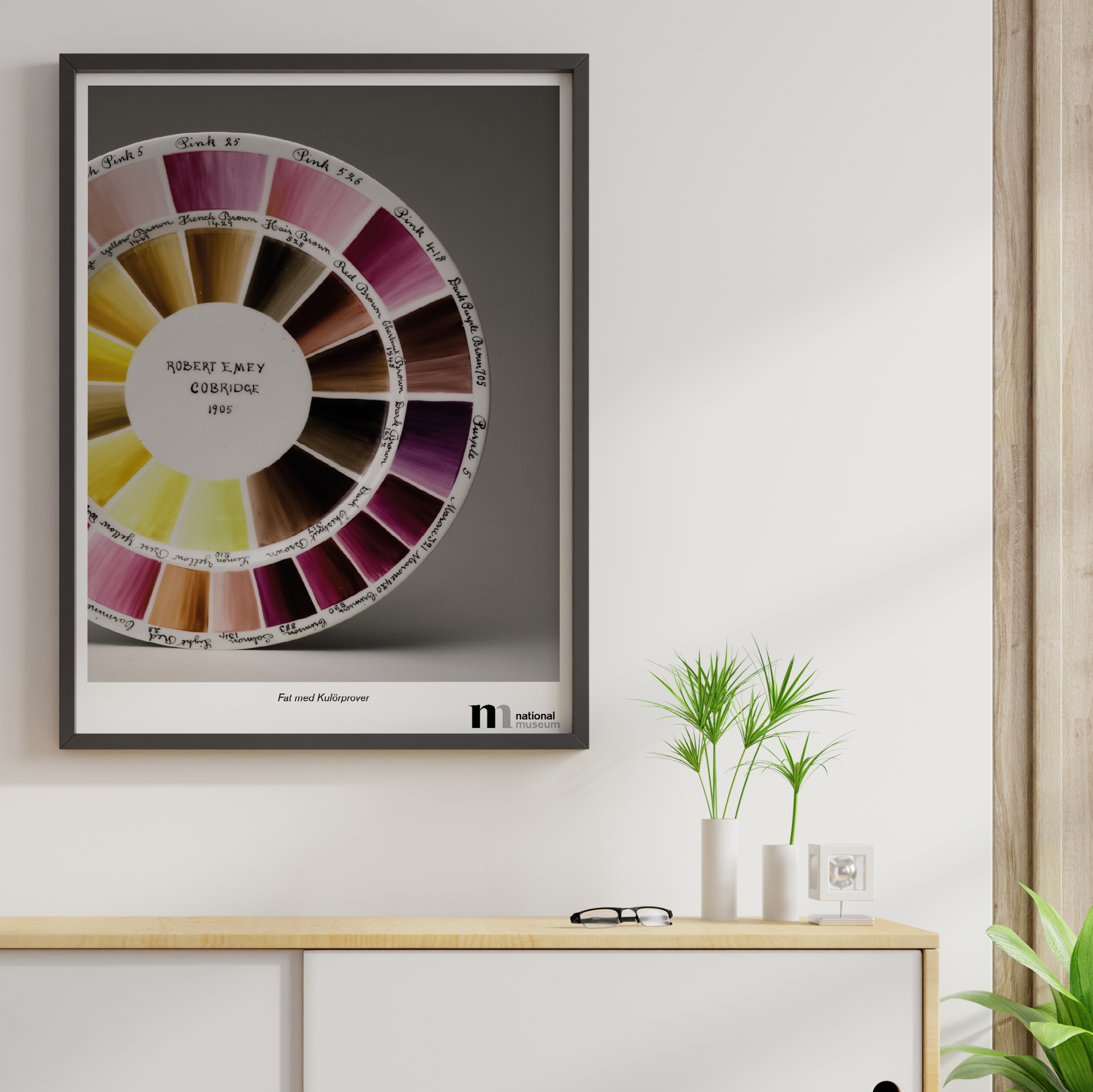 Poster med motiv av kulörprov av rosa och gula färgkombinationer för porslinstillverkning i svart ram över vit bänk