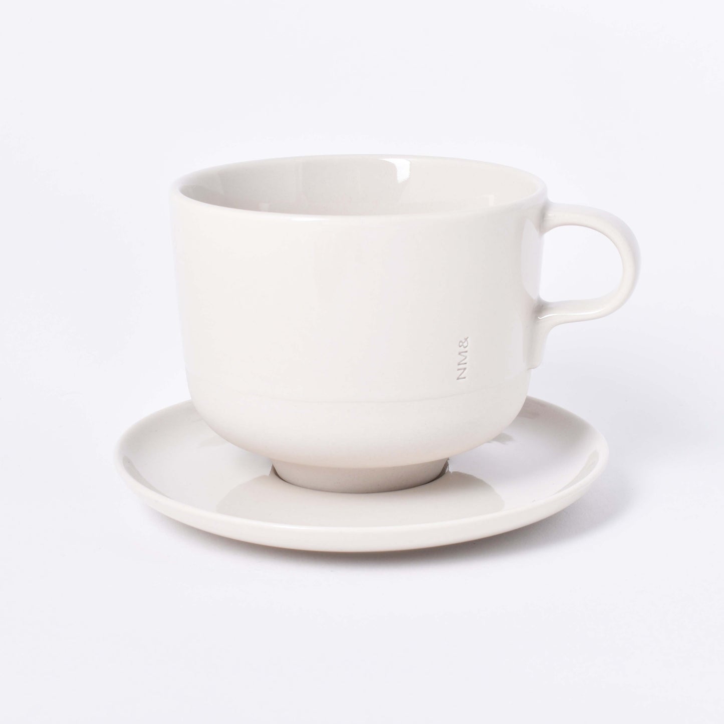 Sandfärgad tekopp med öra och ingraverat NM& monogram på ett runt fat i design av Carina Seth Andersson