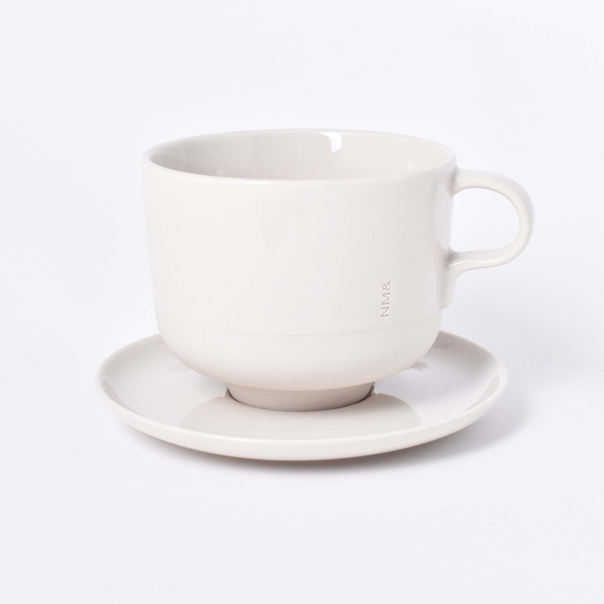 Sandfärgad tekopp med öra och ingraverat NM& monogram på ett runt fat i design av Carina Seth Andersson